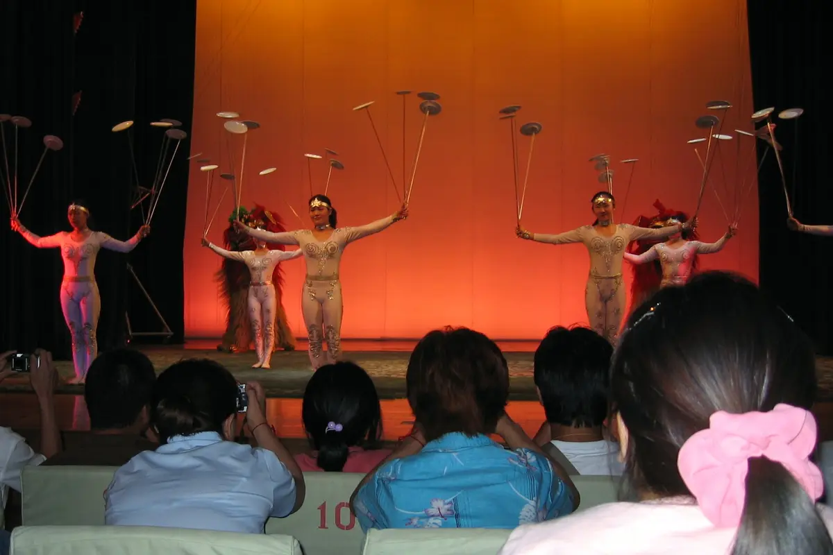 Performers on a stage hold sticks with spinning plates in both hands against an orange backdrop, with the audience's heads in the foreground